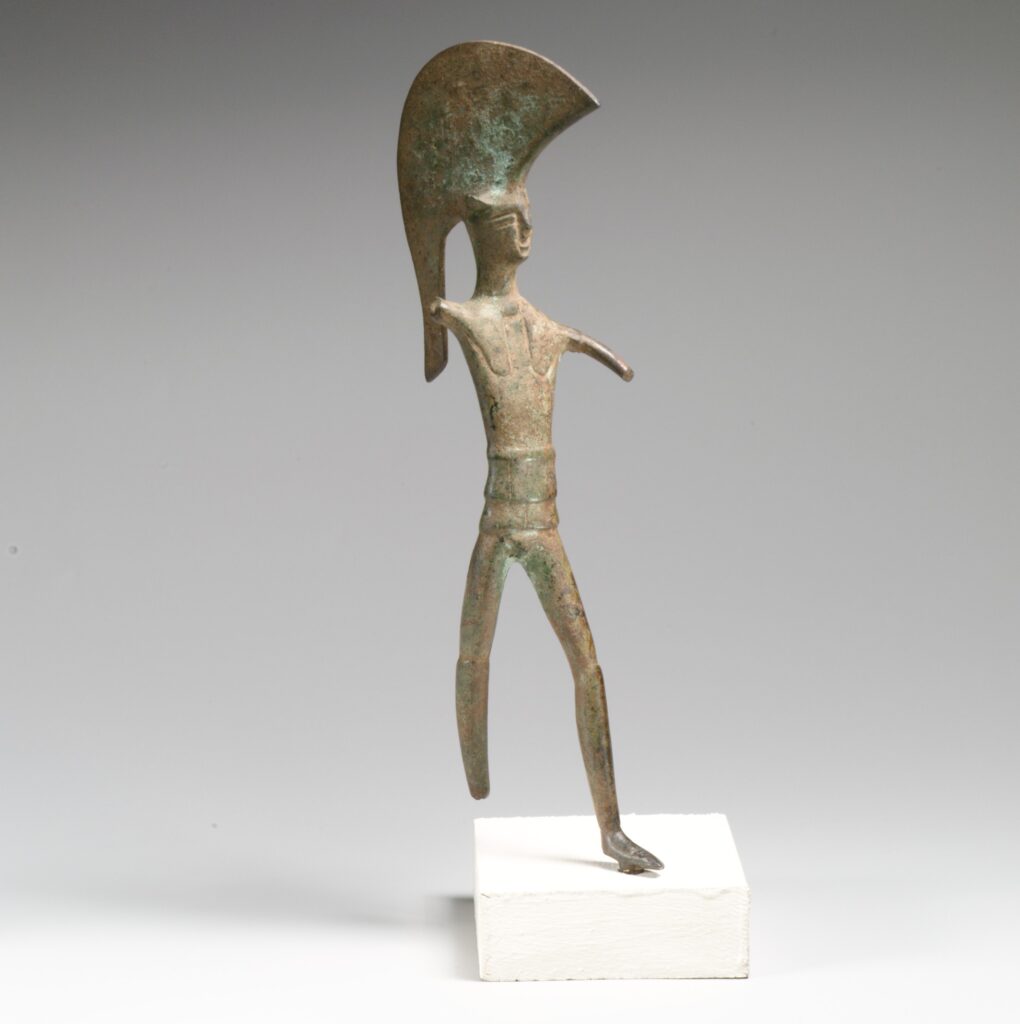 Bronze statue of a warrior wearing a large crested helmet, standing in a rigid pose.